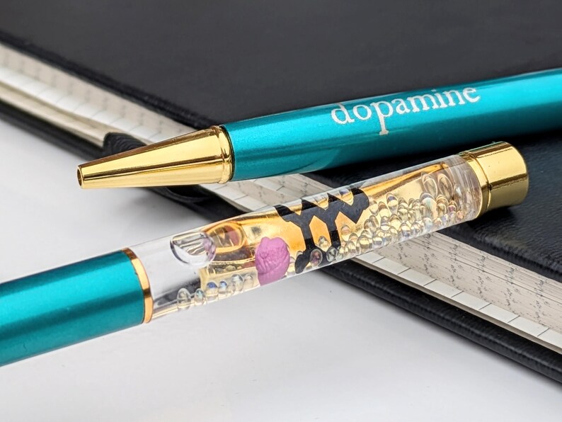 May include: Two teal and gold pens with the word "dopamine" printed on one. One pen has a clear barrel filled with gold liquid, small bubbles, and a pink rose. The pens rest on a black notebook.