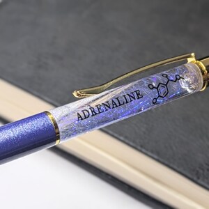 Peut inclure: Un stylo bleu et or avec un corps transparent rempli de paillettes irisées et une structure chimique. Le stylo porte l'inscription "ADRENALINE" sur le corps.