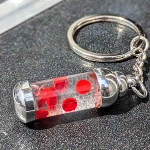 May include: A novelty keychain featuring a clear, cylindrical vial filled with red, circular elements, possibly representing blood cells. The vial has silver-colored metal caps and is attached to a silver keyring.