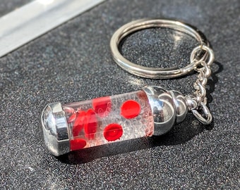 May include: A novelty keyring featuring a clear, cylindrical vial filled with red, circular elements, possibly representing blood cells. The vial has silver-coloured metal caps and is attached to a silver keyring.