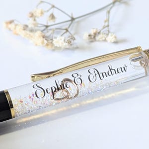 Peut inclure: Un stylo personnalisé avec les noms "Sophie & Andrew" en écriture élégante. Le stylo a un corps transparent rempli de paillettes et de confettis, avec un clip et des accents dorés. La partie inférieure du stylo est noire.