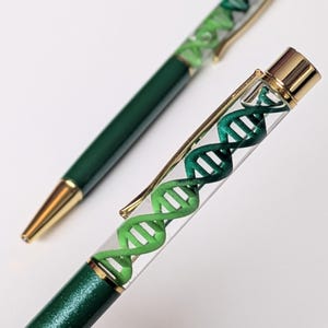 Può includere: Due penne verdi e dorate con un design a elica del DNA. Una penna è a fuoco, mostrando la doppia elica verde e bianca all'interno di un fusto trasparente. Le penne hanno dettagli dorati e un fusto verde scuro.