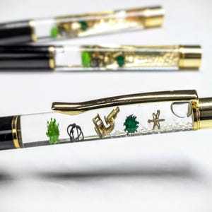 Può includere: Una penna nera e dorata con un corpo trasparente riempito di glitter e piccoli oggetti verdi e dorati. La penna ha una clip dorata e un cappuccio dorato.