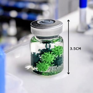 May include: A clear glass vial, 3.5cm tall, with a silver cap, containing green and dark green spherical shapes. The vial is on a white surface, with blurred laboratory equipment in the background.