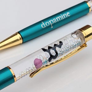 May include: Two teal and gold pens. One pen has the word "dopamine" printed on it. The other pen has a clear barrel filled with small, decorative elements and a gold clip. The pens are on a white surface.