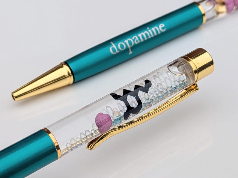 May include: Two teal and gold pens. One pen has the word "dopamine" printed on it. The other pen has a clear barrel filled with small, decorative elements and a gold clip. The pens are on a white surface.
