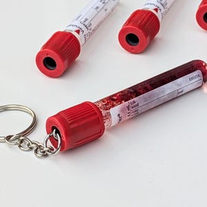 Könnte beinhalten: Ein neuartiger Schlüsselanhänger mit einem Miniatur-Blutröhrchen mit roter Kappe. Das Röhrchen ist mit einer roten Substanz gefüllt und an einem silbernen Schlüsselring befestigt. Im Hintergrund befinden sich mehrere weitere Röhrchen.