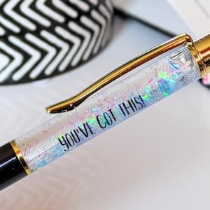 May include: A close-up of a pen with a gold clip and accents. The clear barrel is filled with iridescent glitter and the text "YOU'VE GOT THIS!" is printed on the side. The pen has a black body.