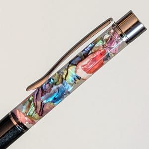 May include: A close-up of a pen with a black barrel and a silver clip. The pen's clear body is filled with colorful, iridescent abalone shell fragments in shades of red, blue, and purple.
