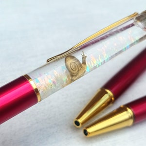 May include: A red and gold pen with a clear barrel filled with iridescent glitter and a gold snail design. The pen is lying on a white surface.