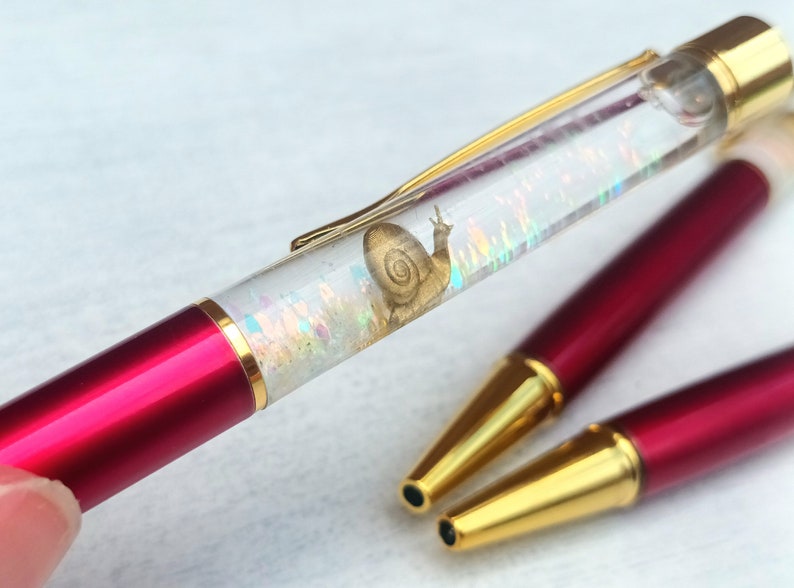 May include: A red and gold pen with a clear barrel filled with iridescent glitter and a gold snail design. The pen is lying on a white surface.