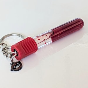 May include: A novelty keychain featuring a miniature, clear vial filled with a deep red liquid, resembling blood. The vial has a red cap and is attached to a silver keyring with a small charm shaped like lips.