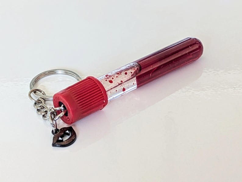 May include: A novelty keychain featuring a miniature, clear vial filled with a deep red liquid, resembling blood. The vial has a red cap and is attached to a silver keyring with a small charm shaped like lips.
