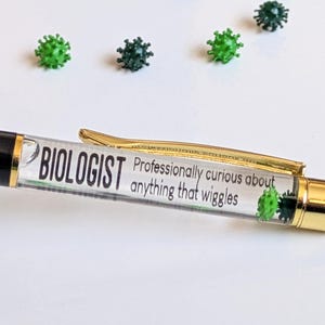 Op de afbeelding: Een zwarte en gouden pen met het woord "BIOLOGIST" en de zin "Professioneel nieuwsgierig naar alles wat beweegt." De pen heeft een doorzichtige schacht met groene, microbe-vormige versieringen. Verschillende groene en donkergroene microbe-vormige objecten zijn rond de pen verspreid.