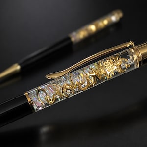 May include: A black ballpoint pen featuring a clear barrel filled with gold-colored star and crescent moon shapes, accented by a gold-toned clip and cap. A second matching pen is visible in the background, slightly out of focus, against a dark surface.