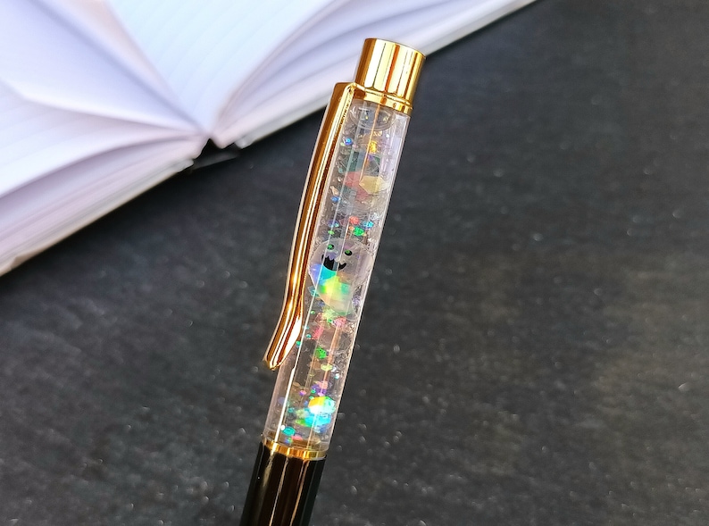 May include: A gold and clear pen with glitter and iridescent flakes inside. The pen has a black cap and a gold clip.