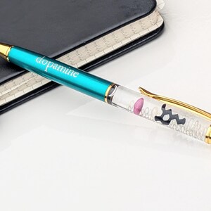 May include: A teal pen with the word "dopamine" printed on the barrel. The pen has gold accents and a clear section filled with small white beads, a pink shape, and a black molecular structure.