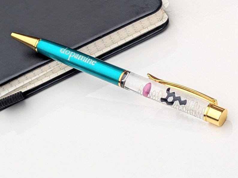 May include: A teal pen with the word "dopamine" printed on the barrel. The pen has gold accents and a clear section filled with small white beads, a pink shape, and a black molecular structure.