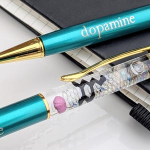 May include: Two teal ballpoint pens with gold accents. One pen has the word "dopamine" printed on it. The other pen has a clear barrel filled with small, colorful objects and a pink pill-shaped object.