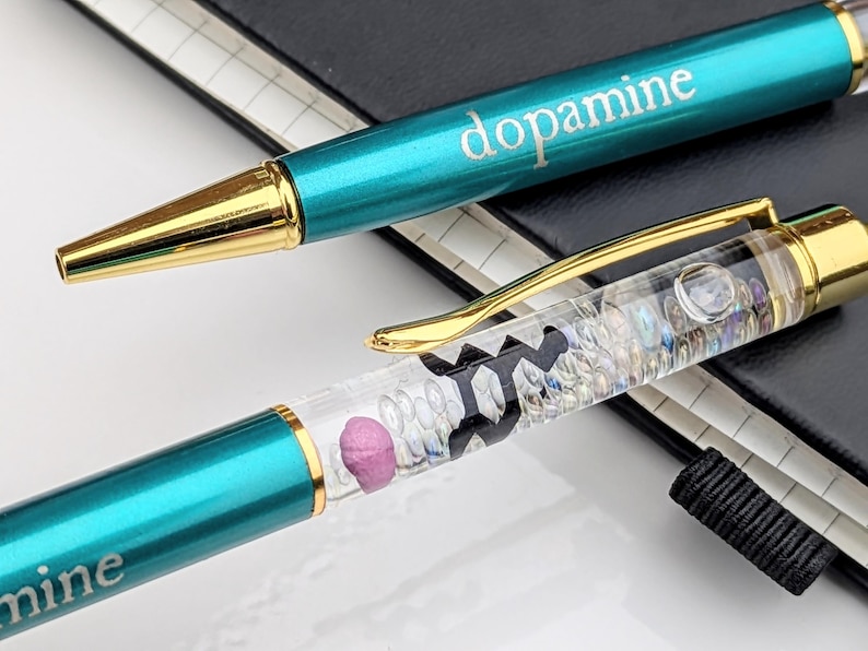 May include: Two teal ballpoint pens with gold accents. One pen has the word "dopamine" printed on it. The other pen has a clear barrel filled with small, colorful objects and a pink pill-shaped object.