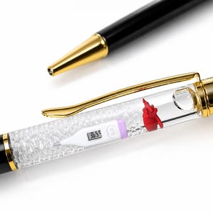 May include: A black ballpoint pen with gold-tone accents featuring a clear barrel filled with small crystals. Inside the barrel, there is a miniature white digital thermometer displaying 36.5 and a small red anatomical heart figurine.