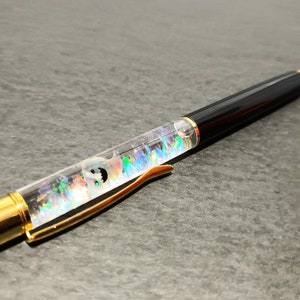 May include: A black and gold pen with a clear barrel filled with colorful glitter and a small, white, iridescent object. The pen has a gold clip and a black ink cartridge.