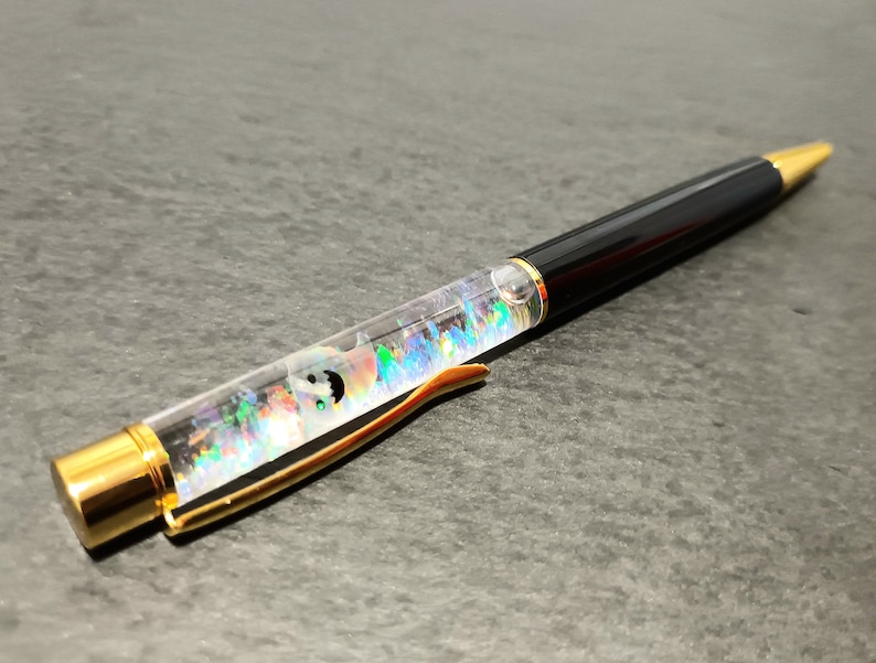May include: A black and gold pen with a clear barrel filled with colorful glitter and a small, white, iridescent object. The pen has a gold clip and a black ink cartridge.