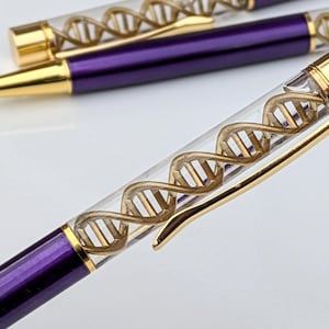 Può includere: Primo piano di una penna viola e oro con un fusto trasparente. All'interno del fusto è presente un design a elica del DNA color oro. La penna ha una clip e una punta dorate e lo sfondo è bianco.