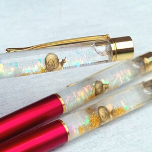 May include: Three clear pens with red and gold accents. Each pen has a small snail charm inside with iridescent glitter.