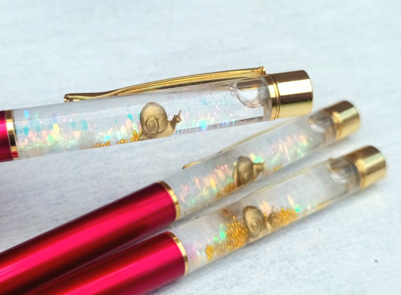 May include: Three clear pens with red and gold accents. Each pen has a small snail charm inside with iridescent glitter.