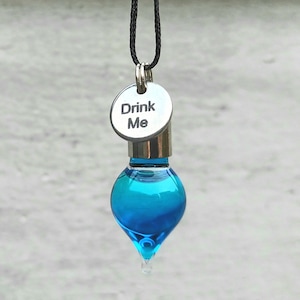 May include: A blue glass bottle pendant with a silver cap and a silver tag that reads "Drink Me". The bottle is shaped like a teardrop and hangs from a black cord.