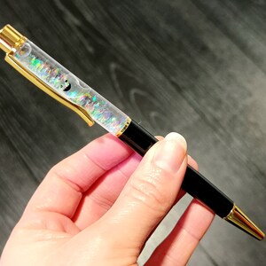 May include: A black and gold pen with a clear barrel filled with iridescent glitter. The glitter is swirling inside the barrel.