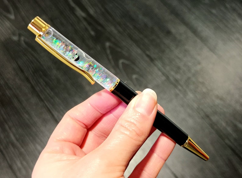 May include: A black and gold pen with a clear barrel filled with iridescent glitter. The glitter is swirling inside the barrel.