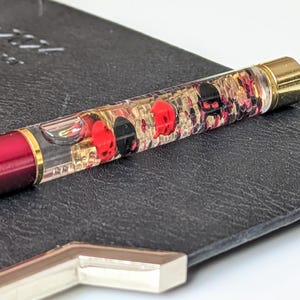 May include: A red pen with a gold-coloured tip and a clear barrel filled with red and black skull designs and gold flakes. The pen rests on a black leather-like surface.