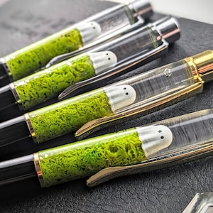 Puede incluir: Primer plano de varios bolígrafos con barriles transparentes, cada uno con una pequeña figura de fantasma blanco y musgo verde. Los bolígrafos tienen cuerpos negros, detalles plateados o dorados y están colocados sobre una superficie oscura.