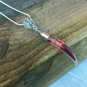May include: A silver necklace with a pendant shaped like a curved horn. The horn is clear with red and white internal marbling. The pendant is attached to a silver chain. The background is a wooden surface.
