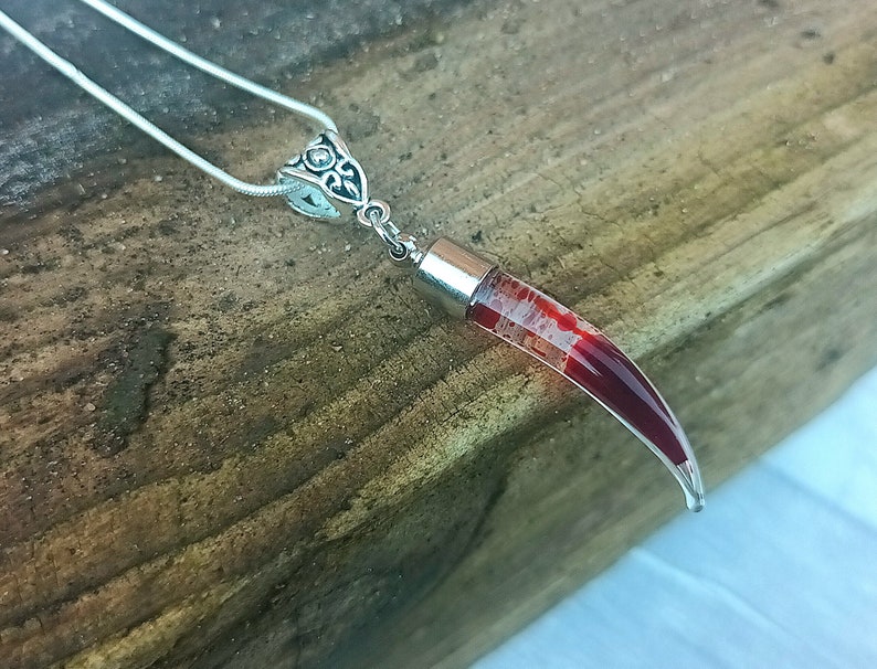 May include: A silver necklace with a pendant shaped like a curved horn. The horn is clear with red and white internal marbling. The pendant is attached to a silver chain. The background is a wooden surface.