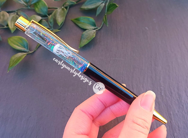 May include: A black and gold pen with a clear barrel filled with iridescent glitter and a white jellyfish design. The pen has the text "carlywarlydesigns" on the side.