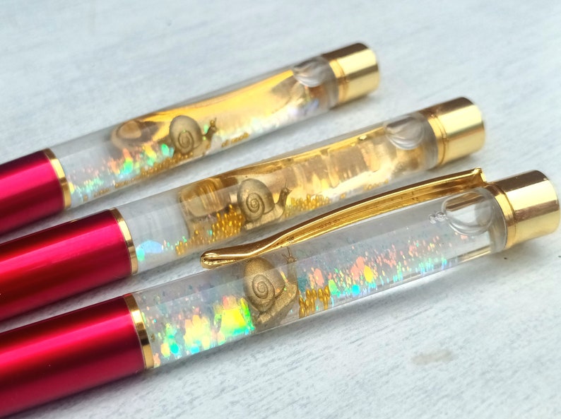 May include: Three red and gold pens with clear barrels filled with iridescent glitter and gold snail charms.