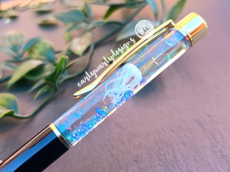 May include: A clear pen with iridescent glitter and a white jellyfish design inside. The pen has a gold-colored clip and a black barrel.