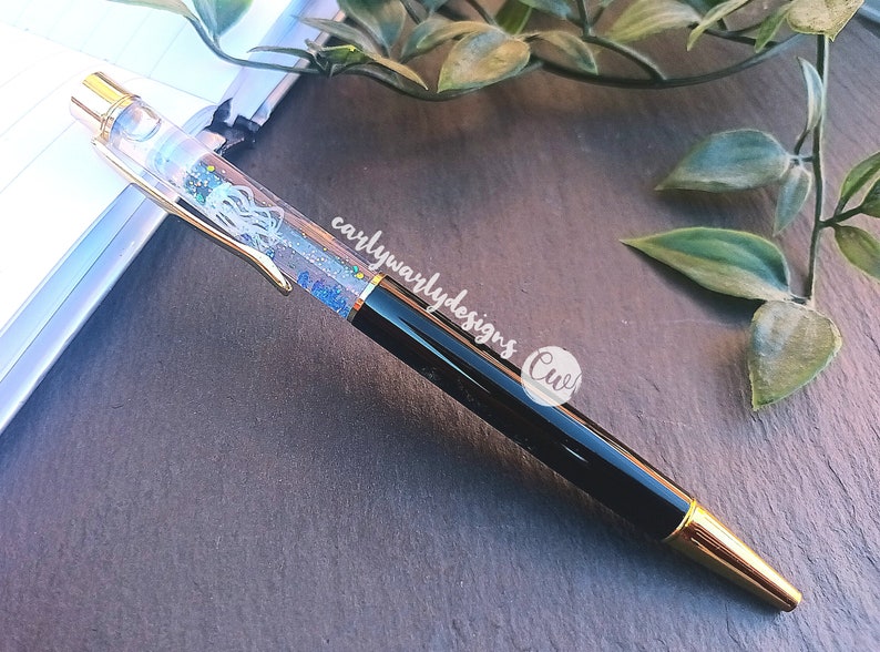 May include: A black and gold pen with a clear barrel filled with blue glitter and a white jellyfish design. The pen has the text "carlywarlydesigns" and a circle with the text "Cwl" on the side.