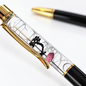 Può includere: Una penna a sfera con fusto trasparente contenente una struttura nera simile a un ramo e una singola foglia rosa. La penna presenta una clip dorata, un tappo dorato e una parte inferiore nera. È visibile una seconda penna nera con punta dorata.