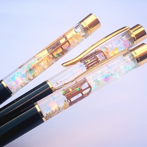 May include: Three gold-capped pens with clear barrels filled with iridescent glitter and a small brown house charm. The pens have black barrels.
