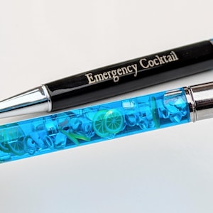 May include: Two pens, one black with "Emergency Cocktail" text, the other with a clear barrel filled with blue liquid, ice-like shapes, and lime slices. Both pens have silver accents.