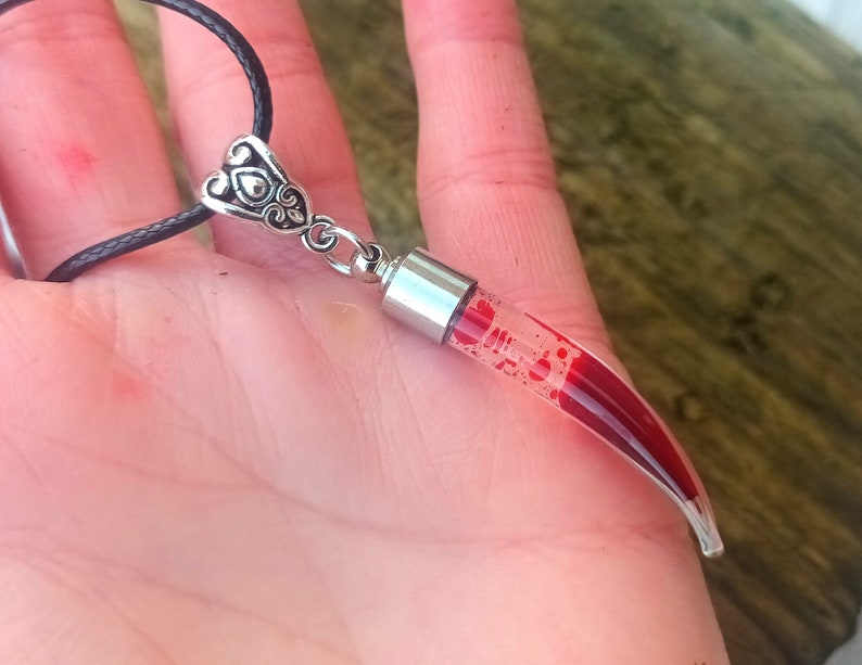 May include: A clear glass pendant filled with red liquid, shaped like a horn or tooth. The pendant hangs from a black cord with a silver-colored decorative bail. The bail has a heart-shaped design.