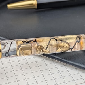 Könnte beinhalten: Ein transparenter Stift mit goldenen Akzenten und einer schwarzen Kappe. Der Stiftschaft enthält Miniatur-Mechanikteile in Gold und ein schwarzes Liniendesign. Im Hintergrund ist ein zweiter Stift mit goldener Spitze zu sehen.