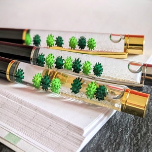 May include: Four black and gold pens with clear barrels filled with green and white beads. The pens are lying on a white notebook.