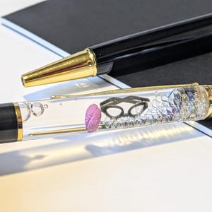 May include: Two black pens with gold accents. One pen has a clear barrel filled with small objects, including a pink brain and black glasses. The other pen is solid black with a gold tip. The pens are on a white surface.