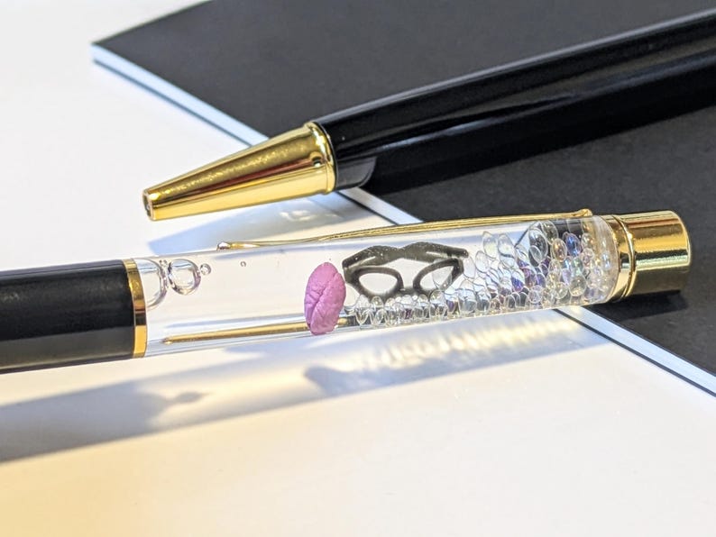 May include: Two black pens with gold accents. One pen has a clear barrel filled with small objects, including a pink brain and black glasses. The other pen is solid black with a gold tip. The pens are on a white surface.