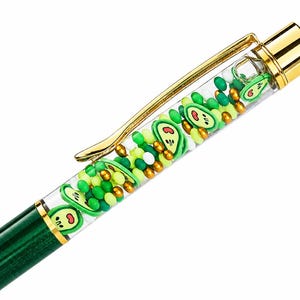 May include: A green pen featuring a clear upper barrel filled with small green, yellow, and white beads along with flat avocado-shaped charms. The pen includes a gold-colored metal clip and a matching gold-colored cap at the top end.